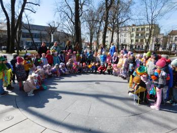 Grupa radosnych dzieci w kolorowych, wiosennych strojach maszeruje ulicami miasta, niosąc kwiaty i dekoracje, świętując pierwszy dzień wiosny