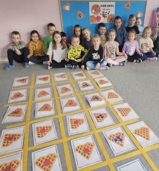 Grupa dzieci siedzi wspólnie za ukończoną planszą, na której widać ułożone karty z goframi i owocami. Wszyscy patrzą w stronę aparatu, uśmiechając się. W tle nadal widoczna jest tablica z dekoracjami z okazji Dnia Gofra.