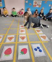 Dziecko układa kartę z obrazkiem gofra na planszy z żółtą siatką, podczas gdy grupa dzieci siedzi w kręgu i obserwuje grę. W tle widać tablicę z napisem „Dzień Gofra 25 marca” oraz dekoracje z goframi.