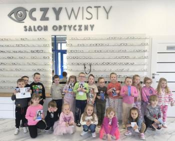 Grupa dzieci pozuje do zdjęcia w salonie optycznym 'Oczywisty'. W tle ściana z ekspozycją okularów, a dzieci trzymają ulotki i materiały informacyjne.
