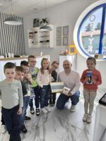 Grupa dzieci wraz z optometrystą w białej koszuli pozuje do zdjęcia w nowoczesnym salonie optycznym. Mężczyzna trzyma w rękach podziękowanie oraz pudełko. 