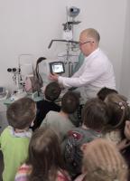 Grupa dzieci obserwuje badanie wzroku w gabinecie optometrycznym. Specjalista w białej koszuli pokazuje im obraz oka na ekranie urządzenia diagnostycznego.
