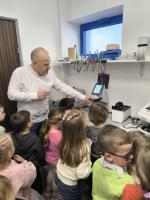 Grupa dzieci obserwuje badanie wzroku w gabinecie optometrycznym. Specjalista w białej koszuli pokazuje im obraz oka na ekranie urządzenia diagnostycznego.