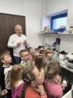 Grupa dzieci obserwuje badanie wzroku w gabinecie optometrycznym. Specjalista w białej koszuli pokazuje im obraz oka na ekranie urządzenia diagnostycznego.