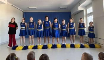 Grupa cheerleaderek w niebieskich strojach z żółtymi pasami stoi na scenie przed publicznością przedszkolaków. W tle znajduje się niebieska dekoracja sceniczna, a sala jest jasno oświetlona dzięki dużym oknom. Publiczność składa się głównie z małych dzieci, które uważnie obserwują występ.