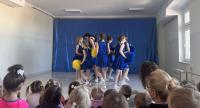 Grupa cheerleaderek w niebieskich strojach z żółtymi pasami stoi na scenie przed publicznością przedszkolaków. W tle znajduje się niebieska dekoracja sceniczna, a sala jest jasno oświetlona dzięki dużym oknom. Publiczność składa się głównie z małych dzieci, które uważnie obserwują występ.