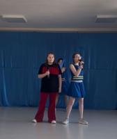 Grupa cheerleaderek w niebieskich strojach z żółtymi pasami stoi na scenie przed publicznością przedszkolaków. W tle znajduje się niebieska dekoracja sceniczna, a sala jest jasno oświetlona dzięki dużym oknom. Publiczność składa się głównie z małych dzieci, które uważnie obserwują występ.
