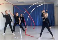 Grupa cheerleaderek w niebieskich strojach z żółtymi pasami stoi na scenie przed publicznością przedszkolaków. W tle znajduje się niebieska dekoracja sceniczna, a sala jest jasno oświetlona dzięki dużym oknom. Publiczność składa się głównie z małych dzieci, które uważnie obserwują występ.