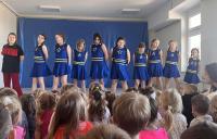 Grupa cheerleaderek w niebieskich strojach z żółtymi pasami stoi na scenie przed publicznością przedszkolaków. W tle znajduje się niebieska dekoracja sceniczna, a sala jest jasno oświetlona dzięki dużym oknom. Publiczność składa się głównie z małych dzieci, które uważnie obserwują występ.
