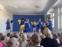 Grupa cheerleaderek w niebieskich strojach z żółtymi pasami stoi na scenie przed publicznością przedszkolaków. W tle znajduje się niebieska dekoracja sceniczna, a sala jest jasno oświetlona dzięki dużym oknom. Publiczność składa się głównie z małych dzieci, które uważnie obserwują występ.