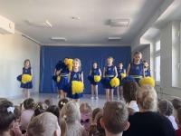 Grupa cheerleaderek w niebieskich strojach z żółtymi pasami stoi na scenie przed publicznością przedszkolaków. W tle znajduje się niebieska dekoracja sceniczna, a sala jest jasno oświetlona dzięki dużym oknom. Publiczność składa się głównie z małych dzieci, które uważnie obserwują występ.