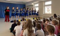 Grupa cheerleaderek w niebieskich strojach z żółtymi pasami stoi na scenie przed publicznością przedszkolaków. W tle znajduje się niebieska dekoracja sceniczna, a sala jest jasno oświetlona dzięki dużym oknom. Publiczność składa się głównie z małych dzieci, które uważnie obserwują występ.