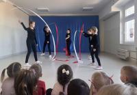 Grupa cheerleaderek w niebieskich strojach z żółtymi pasami stoi na scenie przed publicznością przedszkolaków. W tle znajduje się niebieska dekoracja sceniczna, a sala jest jasno oświetlona dzięki dużym oknom. Publiczność składa się głównie z małych dzieci, które uważnie obserwują występ.