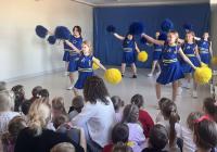 Grupa cheerleaderek w niebieskich strojach z żółtymi pasami stoi na scenie przed publicznością przedszkolaków. W tle znajduje się niebieska dekoracja sceniczna, a sala jest jasno oświetlona dzięki dużym oknom. Publiczność składa się głównie z małych dzieci, które uważnie obserwują występ.