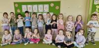 Grupa przedszkolaków pozuje do zdjęcia w sali przedszkolnej, stojąc i siedząc na zielonym dywanie. Dzieci trzymają w rękach papierowe elementy, a za nimi na tablicy znajduje się napis „DZIEŃ GOFRA”, ozdobiony rysunkami gofrów. W tle widać dekoracje ścienne i część wyposażenia sali.
