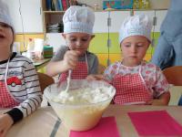 Grupa dzieci w kucharskich czapkach i fartuszkach w przedszkolu siedzi przy długim stole, przygotowując się do pieczenia gofrów z bitą śmietaną. W tle znajdują się kolorowe półki z zabawkami oraz edukacyjne plansze z literami i liczbami.