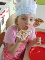 Grupa dzieci w kucharskich czapkach i fartuszkach w przedszkolu siedzi przy długim stole, przygotowując się do pieczenia gofrów z bitą śmietaną. W tle znajdują się kolorowe półki z zabawkami oraz edukacyjne plansze z literami i liczbami.