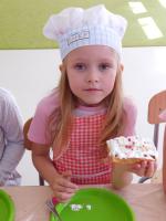Grupa dzieci w kucharskich czapkach i fartuszkach w przedszkolu siedzi przy długim stole, przygotowując się do pieczenia gofrów z bitą śmietaną. W tle znajdują się kolorowe półki z zabawkami oraz edukacyjne plansze z literami i liczbami.
