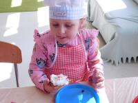 Grupa dzieci w kucharskich czapkach i fartuszkach w przedszkolu siedzi przy długim stole, przygotowując się do pieczenia gofrów z bitą śmietaną. W tle znajdują się kolorowe półki z zabawkami oraz edukacyjne plansze z literami i liczbami.
