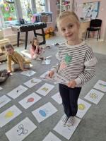 Grupa dzieci siedzących w kole na dywanie w przedszkolu, wokół kart z rysunkami różnych przedmiotów, zwierząt i symboli. W centrum znajduje się ścieżka z kart prowadząca od oznaczonego miejsca 'START'. W tle widać kolorowe meble, zabawki i dekoracje na ścianach.