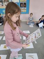 Grupa dzieci siedzących w kole na dywanie w przedszkolu, wokół kart z rysunkami różnych przedmiotów, zwierząt i symboli. W centrum znajduje się ścieżka z kart prowadząca od oznaczonego miejsca 'START'. W tle widać kolorowe meble, zabawki i dekoracje na ścianach.