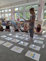 Grupa dzieci siedzących w kole na dywanie w przedszkolu, wokół kart z rysunkami różnych przedmiotów, zwierząt i symboli. W centrum znajduje się ścieżka z kart prowadząca od oznaczonego miejsca 'START'. W tle widać kolorowe meble, zabawki i dekoracje na ścianach.