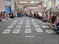 Grupa dzieci siedzących w kole na dywanie w przedszkolu, wokół kart z rysunkami różnych przedmiotów, zwierząt i symboli. W centrum znajduje się ścieżka z kart prowadząca od oznaczonego miejsca 'START'. W tle widać kolorowe meble, zabawki i dekoracje na ścianach.