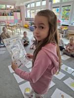 Grupa dzieci siedzących w kole na dywanie w przedszkolu, wokół kart z rysunkami różnych przedmiotów, zwierząt i symboli. W centrum znajduje się ścieżka z kart prowadząca od oznaczonego miejsca 'START'. W tle widać kolorowe meble, zabawki i dekoracje na ścianach.