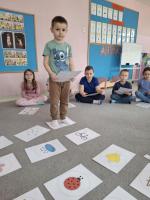 Grupa dzieci siedzących w kole na dywanie w przedszkolu, wokół kart z rysunkami różnych przedmiotów, zwierząt i symboli. W centrum znajduje się ścieżka z kart prowadząca od oznaczonego miejsca 'START'. W tle widać kolorowe meble, zabawki i dekoracje na ścianach.