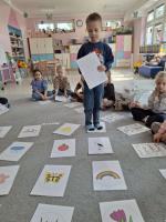 Grupa dzieci siedzących w kole na dywanie w przedszkolu, wokół kart z rysunkami różnych przedmiotów, zwierząt i symboli. W centrum znajduje się ścieżka z kart prowadząca od oznaczonego miejsca 'START'. W tle widać kolorowe meble, zabawki i dekoracje na ścianach.