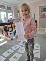 Grupa dzieci siedzących w kole na dywanie w przedszkolu, wokół kart z rysunkami różnych przedmiotów, zwierząt i symboli. W centrum znajduje się ścieżka z kart prowadząca od oznaczonego miejsca 'START'. W tle widać kolorowe meble, zabawki i dekoracje na ścianach.