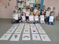 Grupa dzieci siedzących w kole na dywanie w przedszkolu, wokół kart z rysunkami różnych przedmiotów, zwierząt i symboli. W centrum znajduje się ścieżka z kart prowadząca od oznaczonego miejsca 'START'. W tle widać kolorowe meble, zabawki i dekoracje na ścianach.