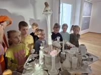 Grupa dzieci ogląda wystawę w muzeum. Dzieci są skupione i zaciekawione eksponatami. W tle widoczne są jasne ściany galerii oraz oprawione dokumenty i fotografie.