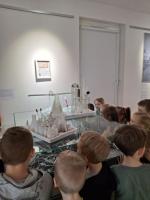 Grupa dzieci ogląda wystawę w muzeum. Dzieci są skupione i zaciekawione eksponatami. W tle widoczne są jasne ściany galerii oraz oprawione dokumenty i fotografie.