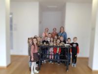 Grupa dzieci ogląda wystawę w muzeum. Dzieci są skupione i zaciekawione eksponatami. W tle widoczne są jasne ściany galerii oraz oprawione dokumenty i fotografie.