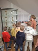 Grupa dzieci ogląda wystawę w muzeum. Dzieci są skupione i zaciekawione eksponatami. W tle widoczne są jasne ściany galerii oraz oprawione dokumenty i fotografie.