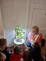 Grupa dzieci ogląda wystawę w muzeum. Dzieci są skupione i zaciekawione eksponatami. W tle widoczne są jasne ściany galerii oraz oprawione dokumenty i fotografie.