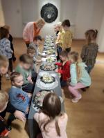 Grupa dzieci ogląda wystawę w muzeum. Dzieci są skupione i zaciekawione eksponatami. W tle widoczne są jasne ściany galerii oraz oprawione dokumenty i fotografie.