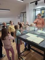 Grupa dzieci ogląda wystawę w muzeum. Dzieci są skupione i zaciekawione eksponatami. W tle widoczne są jasne ściany galerii oraz oprawione dokumenty i fotografie.