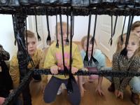 Grupa dzieci ogląda wystawę w muzeum. Dzieci są skupione i zaciekawione eksponatami. W tle widoczne są jasne ściany galerii oraz oprawione dokumenty i fotografie.