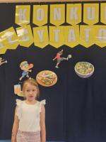Dzieci biorące udział w przedszkolnym konkursie recytatorskim – stoją na scenie, recytują wiersze lub odbierają dyplomy i nagrody. W tle widać dekoracje tematyczne i jury.